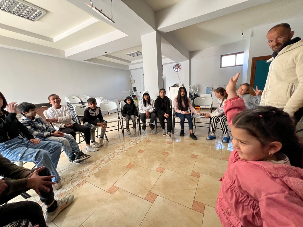 An illustrated, smiling woman sits at a desk in front of a laptop and a yuppon flower plant, wearing an orange top and bro...
